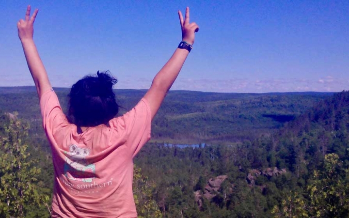A person lifts two peace signs into the air while looking out over a vast, green wooded area. 